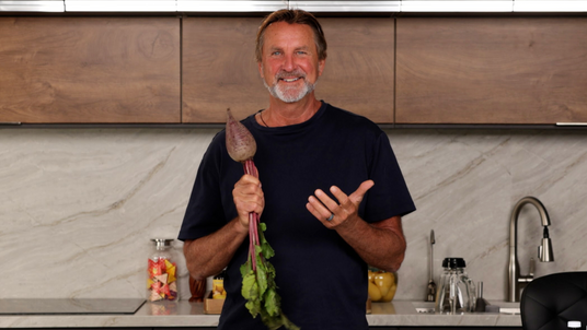 Man in a kitchen holding a bunch of greens, with bottles and kitchen items in the background.